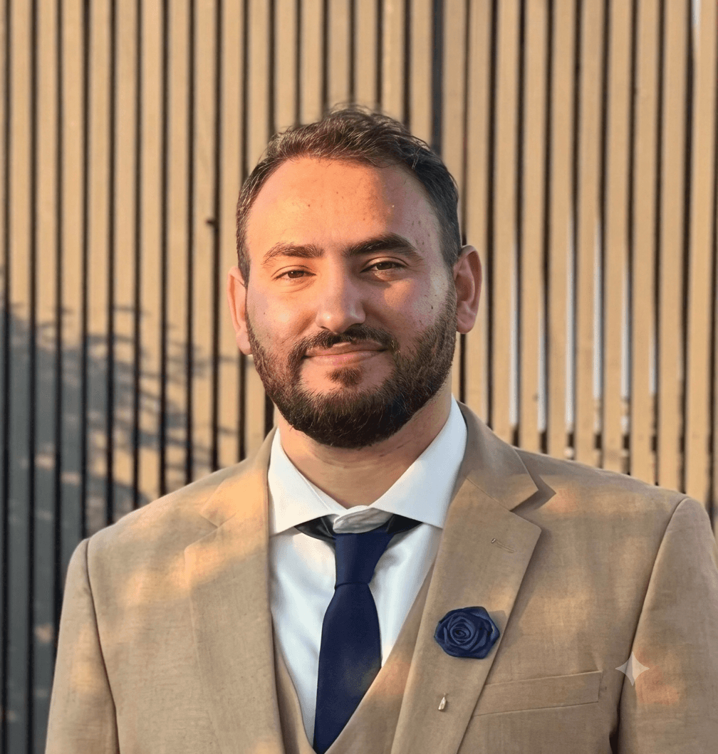 Bearded man in a tan suit and blue tie smiling against a vertical wooden slat fence.
